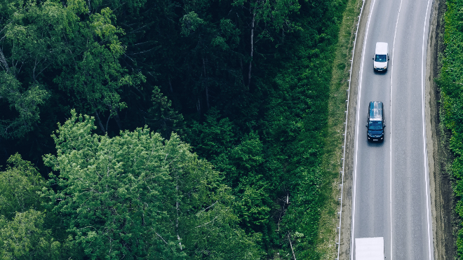 White van driving behind a black van along a single carriageway road beside a forest.